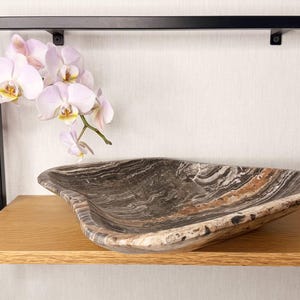 Könnte beinhalten: Eine dekorative, wellenförmige Steinschale mit brauner, grauer und weißer Marmorierung steht auf einem Holzregal. Ein schwarzer Metallrahmen und ein Zweig Orchideen mit rosa und gelben Blüten sind ebenfalls zu sehen.