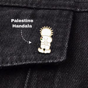Pode incluir: Um alfinete de esmalte branco com uma figura de desenho animado com um halo semelhante ao sol, segurando uma pequena bandeira palestina. O alfinete está rotulado "Palestine Handala" em texto branco. O alfinete está preso a uma jaqueta jeans preta.