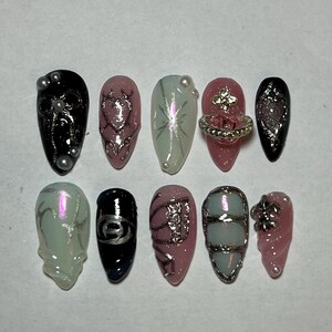 Puede incluir: Diez uñas postizas diferentes con varios diseños. Los diseños incluyen un corazón, una estrella, una corona, un patrón geométrico y un remolino. Las uñas son en tonos de rosa, blanco y negro.
