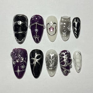 Puede incluir: Conjunto de diez uñas postizas con varios diseños. Las uñas son de forma ovalada y presentan una mezcla de colores negro, blanco y morado. Algunas uñas tienen detalles plateados, incluyendo cadenas, estrellas y lazos.