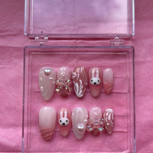 Puede incluir: Un conjunto de diez uñas postizas de color rosa y blanco con diseños de conejitos y lazos. Las uñas se muestran en una caja de plástico transparente.