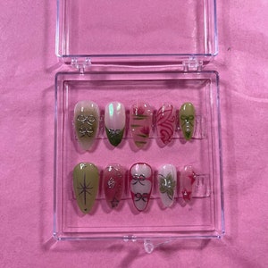 Puede incluir: Una caja de plástico transparente que contiene diez uñas postizas con varios diseños. Los diseños incluyen una estrella, un lazo, flores y una mariposa. Las uñas son de color rosa claro y verde.