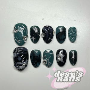 May include: A collection of press-on nails in shades of dark teal, black, and silver. Some nails are adorned with stars and gems, while others feature abstract patterns. The image includes the text "desy's nails".