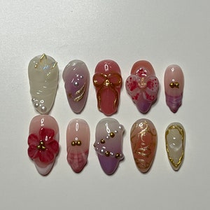 Puede incluir: Diez diseños de uñas diferentes que presentan varios colores, patrones y adornos. Los diseños incluyen una uña blanca con un remolino dorado, una uña rosa con un lazo dorado, una uña morada con cuentas doradas y una uña rosa con una flor roja.