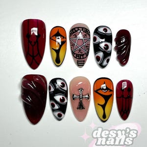 May include: A set of ten press-on nails with various designs. The nails feature a range of colors, including red, black, orange, and pink. Designs include abstract shapes, eye motifs, a pentagram, and a cross. The brand name "desy's nails" is visible.