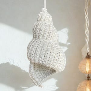 May include: A cream-coloured, crocheted seashell-shaped lamp hanging from a white cord. The lamp has a spiral design and a textured surface. The background is a white wall, with a glimpse of other hanging lamps in the distance.