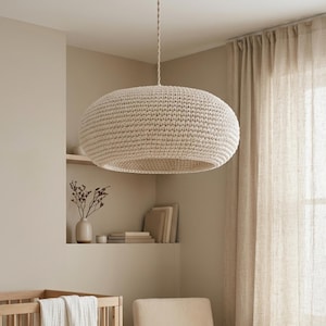 Puede incluir: Un cuarto de niños de tonos neutros con una cuna de madera, una mecedora y una gran lámpara colgante tejida. La habitación cuenta con cortinas beige y una alfombra texturizada, creando un espacio tranquilo y acogedor. Una manta blanca está sobre la cuna.