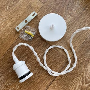 May include: Overhead shot of a white pendant light kit. The kit includes a white ceiling canopy, a white lamp holder with a black band, a white braided cord, a mounting bracket, and screws. The components are arranged on a wooden surface.