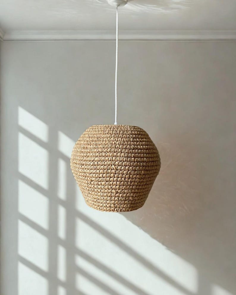 May include: A beige, woven pendant light fixture. The light has a rounded, geometric shape and is suspended from a white cord attached to a ceiling fixture. The light casts shadows on the wall.
