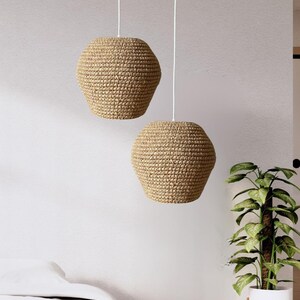 May include: Two pendant lights with woven, beige shades hang from white cords. The shades have a hexagonal shape and a textured surface. The lights are suspended in a room with a white wall and a potted plant.