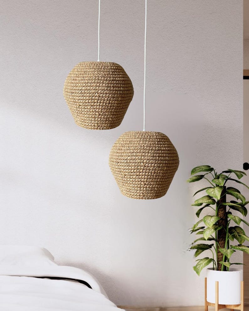 May include: Two pendant lights with woven, beige shades hang from white cords. The shades have a hexagonal shape and a textured surface. The lights are suspended in a room with a white wall and a potted plant.