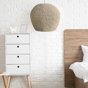 May include: A beige, spherical pendant light hangs from the ceiling in a bedroom. The light fixture has a textured, woven appearance. A white dresser with black drawer pulls and a wooden headboard are also visible.