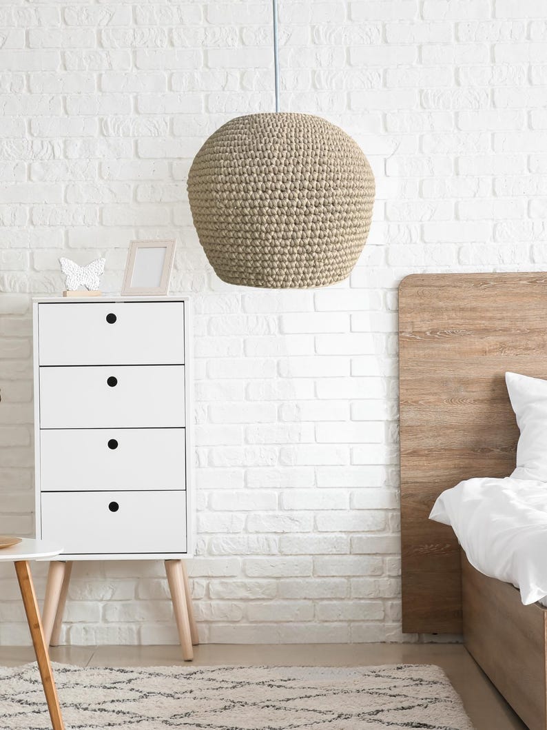 May include: A beige, spherical pendant light hangs from the ceiling in a bedroom. The light fixture has a textured, woven appearance. A white dresser with black drawer pulls and a wooden headboard are also visible.