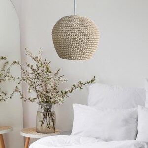 May include: A beige, spherical pendant light made of woven material hangs from a white cord. The light fixture is the focal point, with a soft, textured appearance. The background includes a bed with white linens and a vase of flowers.