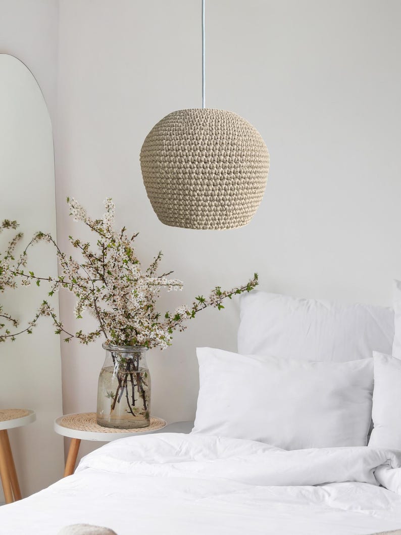 May include: A beige, spherical pendant light made of woven material hangs from a white cord. The light fixture is the focal point, with a soft, textured appearance. The background includes a bed with white linens and a vase of flowers.