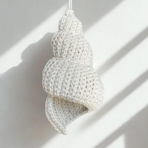 Könnte beinhalten: Gehäkeltes Muschelschmuckstück in Weiß- und Grautönen, an einem dünnen weißen Faden aufgehängt. Das Spiraldesign ist detailliert, und die Textur ist sichtbar. Der Hintergrund ist eine schlichte weiße Oberfläche mit Schatten.