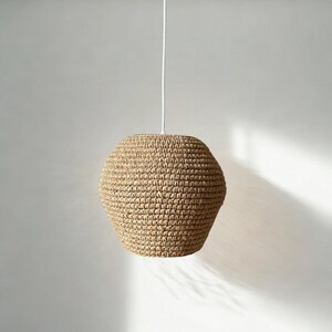 May include: A woven pendant light fixture with a natural, beige-colored shade. The shade has a rounded, geometric shape and hangs from a white cord and ceiling mount. The light fixture is suspended against a plain white background.