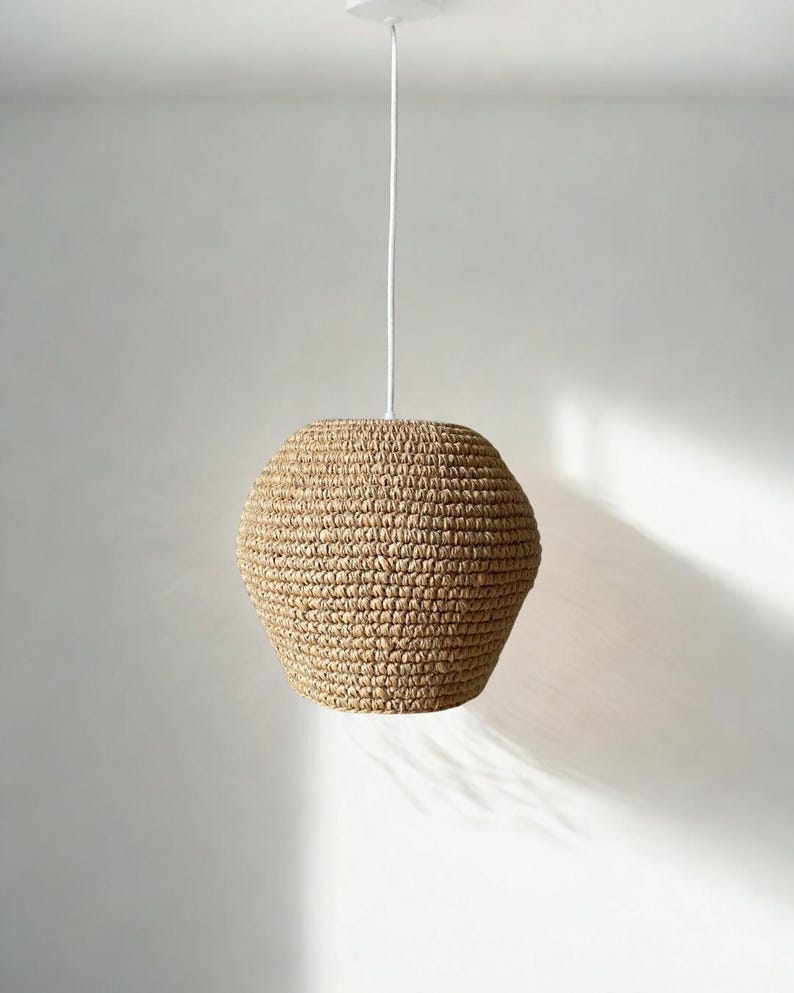 May include: A woven pendant light fixture with a natural, beige-colored shade. The shade has a rounded, geometric shape and hangs from a white cord and ceiling mount. The light fixture is suspended against a plain white background.