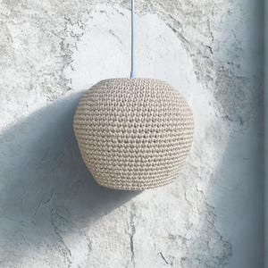 May include: A beige, spherical pendant light with a crocheted texture. The light hangs from a pale blue cord against a textured, off-white wall. The light fixture is approximately 10 inches in diameter.
