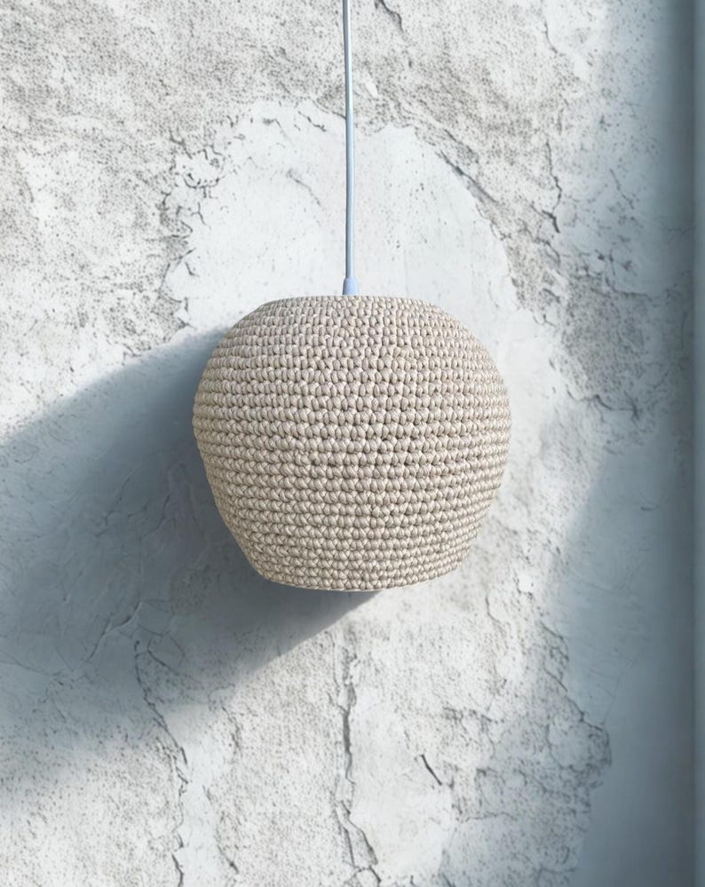 May include: A beige, spherical pendant light with a crocheted texture. The light hangs from a pale blue cord against a textured, off-white wall. The light fixture is approximately 10 inches in diameter.