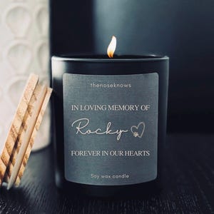 May include: A black soy wax candle with a lit flame. The label reads "IN LOVING MEMORY OF Rocky FOREVER IN OUR HEARTS." The candle has a wooden lid and the text "thenoseknows" is at the top of the label.