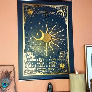 Op de afbeelding: Een zwarte en gouden poster met de tekst "Calendrier Lunaire 2025" en een gedetailleerde illustratie van de maanfasen gedurende het jaar.