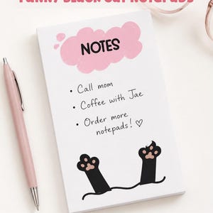 May include: A rectangular notepad featuring a pink cloud graphic with the text NOTES, a handwritten to-do list, and two black cat paws at the bottom. The notepad is accompanied by a pink pen and pink-rimmed glasses, with text reading FUNNY BLACK CAT NOTEPADS and DIGITAL DOWNLOAD.