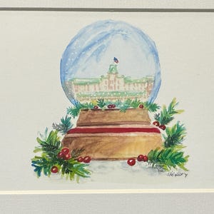May include: Watercolor painting of a snow globe featuring a building with a flag inside a blue globe. The base is brown with a red stripe, surrounded by green sprigs and red berries. The artwork is framed in white.