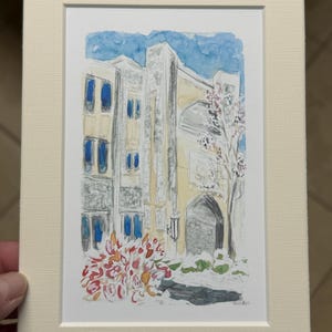 May include: Watercolor painting of a building with blue windows and a stone archway, framed in a cream-colored mat. A tree with pink blossoms and red and orange bushes are in the foreground. The sky is blue.