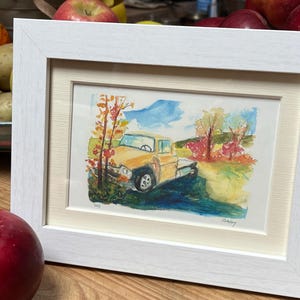 May include: A framed watercolor painting of a yellow truck driving through a fall landscape. The truck is driving on a dirt road with trees on either side. The trees are red, orange, and yellow. The sky is blue with white clouds. The painting is signed "Shay" in the bottom left corner and "Hadley" in the bottom right corner.
