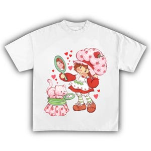 Puede incluir: Camiseta blanca con una ilustraci&oacute;n de dibujos animados de un personaje con un sombrero de fresa, sosteniendo un espejo. Un gato rosa est&aacute; sentado en un recipiente rosa y verde. Corazones rojos est&aacute;n dispersos alrededor de la imagen. El personaje lleva un vestido rojo y blanco.