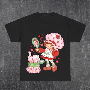 Puede incluir: Camiseta negra con una ilustraci&oacute;n de dibujos animados de una ni&ntilde;a con un sombrero de fresa, sosteniendo un espejo. Un gato rosa est&aacute; sentado en un pedestal con tem&aacute;tica de fresa. Corazones rojos rodean a los personajes. La ni&ntilde;a lleva un vestido rojo y blanco.