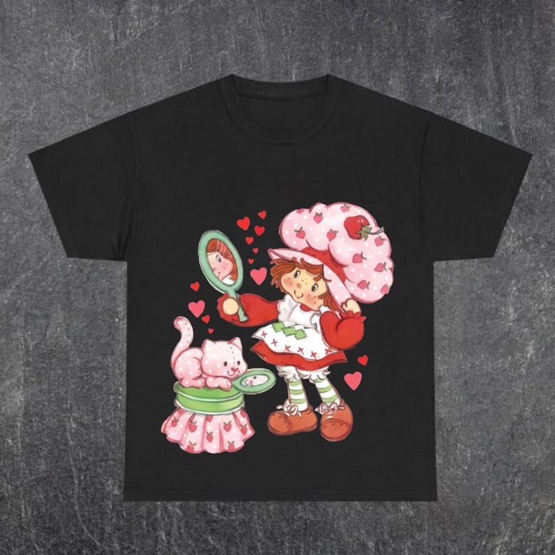 Puede incluir: Camiseta negra con una ilustraci&oacute;n de dibujos animados de una ni&ntilde;a con un sombrero de fresa, sosteniendo un espejo. Un gato rosa est&aacute; sentado en un pedestal con tem&aacute;tica de fresa. Corazones rojos rodean a los personajes. La ni&ntilde;a lleva un vestido rojo y blanco.