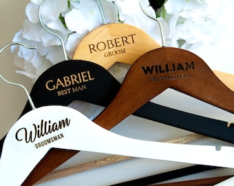 May include: Four wooden hangers with engraved names and titles, including "Robert Groom", "Gabriel Best Man", and two "William Groomsman". The hangers are in different colors: natural wood, black, brown, and white.