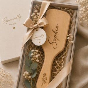 Può includere: Una spazzola per capelli in legno chiaro incisa con il nome Sophia e un piccolo rametto floreale, presentata in una scatola regalo trasparente con un nastro beige, un'etichetta rotonda con la scritta Especially for you ed eucalipto.