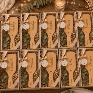 Può includere: Dieci scatole regalo rettangolari contenenti ciascuna una spazzola per capelli in legno chiaro incisa con un nome unico, tra cui Ashley, Hailey, Sophia, Olivia, Emma, Charlotte, Amelia, Isabella, Mia e Harper, decorate con nastro e verde.
