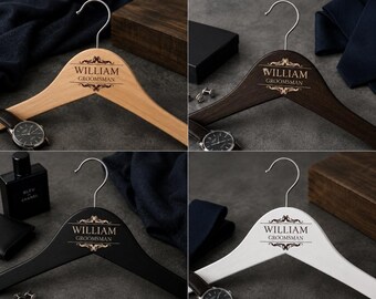 May include: Four wooden clothes hangers in different colors: natural, dark brown, black, and white. Each hanger has a silver hook and is engraved with the name "WILLIAM" and the word "GROOMSMAN" in a decorative font. The hangers are displayed with accessories.