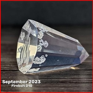 May include: A clear, faceted ten-sided die, known as a D10, featuring a flame icon on the top face and engraved numbers. The text "REVEL BROKER" is visible on the side, with "September 2023" and "Firebolt D10" printed at the bottom of the image.