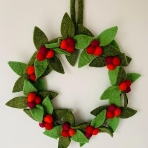 May include: A felt wreath with green leaves and red berry clusters. The wreath is circular and has a green loop for hanging. The design is simple and festive, suitable for seasonal decoration.