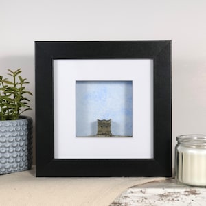 May include: A framed 3D artwork featuring a small, detailed model of a building against a blue and white background. The frame is black with a white mount.