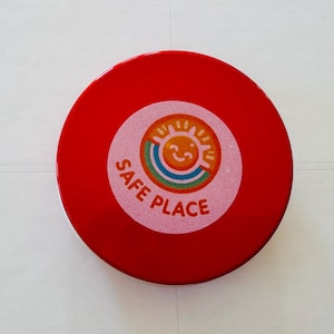 May include: A circular, red object with a central sticker. The sticker has a smiling sun, rainbow, and the words "SAFE PLACE". The object has a glossy finish. The sticker is white and pink.