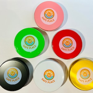 Może przedstawiać: Sześć okrągłych metalowych puszek z pokrywkami, każda w innym kolorze i z grafiką uśmiechniętego słońca z tekstem "SAFE PLACE".
