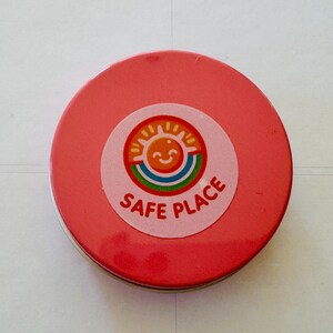 May include: A circular, red tin with a sticker on the lid. The sticker shows a smiling sun above a rainbow, with the words "SAFE PLACE". The tin is probably for storage or as a container.