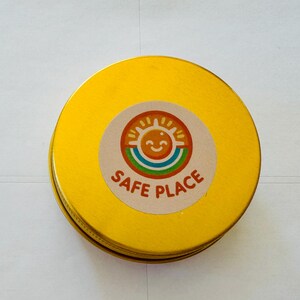 May include: A circular, yellow tin with a lid. A round sticker on top displays a smiling sun, rainbow, and the words "SAFE PLACE" in brown. The tin is on a white surface.