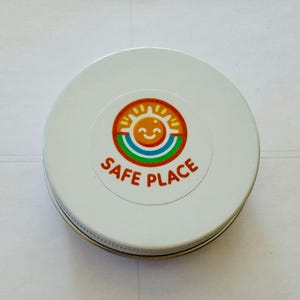 Może przedstawiać: Biała, okrągła puszka z kolorową naklejką na wieczku. Naklejka przedstawia uśmiechnięte słońce, tęczę i czerwony napis "SAFE PLACE". Puszka ma srebrną obwódkę.