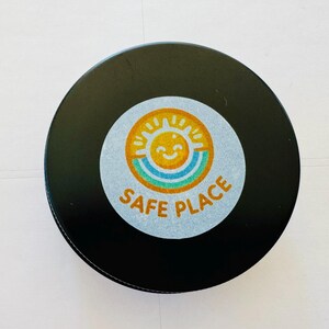 May include: A black, round coaster with a circular sticker in the center. The sticker features a smiling sun with a rainbow and the words "SAFE PLACE" in orange text. The coaster is likely made of a durable material.