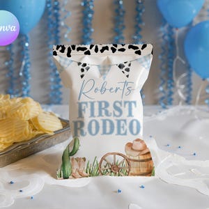 May include: A white chip bag with the text "Robert's FIRST RODEO" in light blue. The bag has a cow print design at the top and a watercolor illustration of a cactus, cowboy boots, a barrel, and a wagon wheel. Blue balloons are in the background.