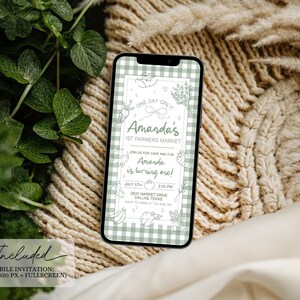 May include: A smartphone displays a mobile invitation for Amanda's 1st Farmers Market and birthday celebration. The invitation features a green and white gingham pattern, with text and illustrations of vegetables and fruits. The background includes greenery and a beige knitted texture.