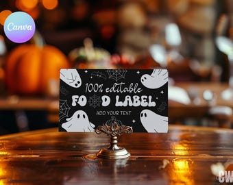 May include: A black Halloween food label with white ghost illustrations, spiderwebs, and the text "100% editable FOOD LABEL ADD YOUR TEXT." The label is held by a decorative gold stand. A pumpkin is in the background.