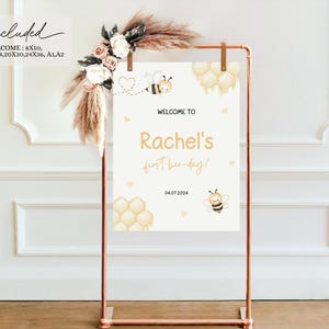 May include: A white welcome sign with a gold honeycomb design and a cute bee illustration. The sign says "Welcome to Rachel's First Bee Day!" and includes the date "04.07.2024".